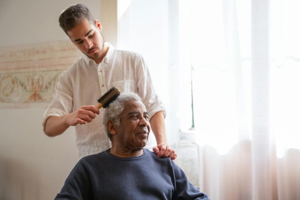 Young man gently combs elderly man's hair by a sunlit window, portraying caregiving and family bond.